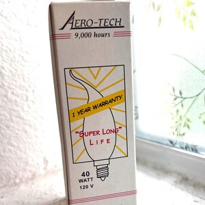 May include: A rectangular white cardboard box for an Aero-Tech light bulb. The box features a graphic of a light bulb with yellow rays, text reading 9,000 hours, 1 YEAR WARRANTY, SUPER LONG LIFE, 40 WATT, 120 V, and A USA company in red and black.