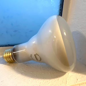 May include: A large, pear-shaped light bulb with a white and tan finish. The bulb has a gold-colored base and is resting on a white surface. The text "620 Lumens" is visible on the bulb.