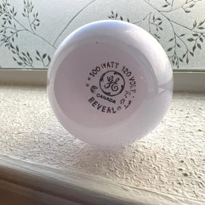 Pode incluir: Uma lâmpada GE Reveal branca, 100 watts, 120 volts. A lâmpada tem formato redondo e está marcada com "Canadá". A lâmpada está sobre uma superfície branca.