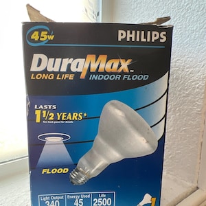 May include: A Philips DuraMax indoor flood light bulb in a box. The box is black and blue with the text "DuraMax" and "Indoor Flood". The bulb is 45 watts and lasts 1 1/2 years. The light output is 340 lumens.