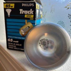 May include: A silver Philips reflector spot light bulb with the text "PHILIPS" and "U.S.A." printed on the bulb. The bulb is next to a black and blue box that reads "PHILIPS Track SPOT 120V 65w".