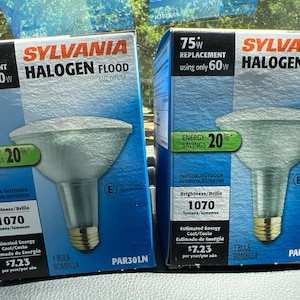 Puede incluir: Dos cajas de bombillas Sylvania Halogen Flood. Cada caja azul presenta un gráfico de la bombilla, indicando reemplazo de 75W usando solo 60W, y un ahorro de energía del 20%. Las cajas también muestran el brillo de 1070 lúmenes.