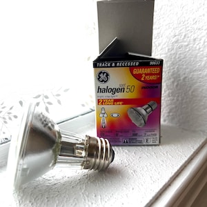 Puede incluir: Una bombilla halógena de 50 puntos plateada con una caja que dice "¡GARANTIZADO 2 AÑOS!" y "INTERIOR". La caja es negra, roja y amarilla. La bombilla está sobre una superficie blanca.