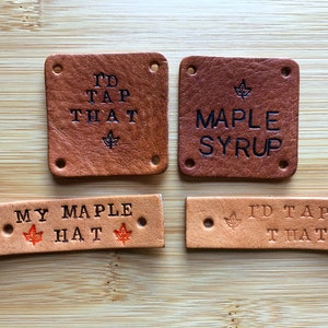 May include: Four leather patches with different maple syrup themed sayings. The patches are brown and tan with black and red lettering. The sayings are "I'd Tap That", "Maple Syrup", "My Maple Hat", and "I'd Tap That".