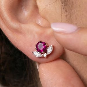 Puede incluir: Primer plano de un pendiente de botón con una gema facetada de color rojo intenso en el centro, rodeada de piedras más pequeñas, blancas, con forma de marquesa. El pendiente está engastado en un metal dorado y se lleva en una oreja.