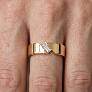 Op de afbeelding: Een gepolijste gouden ring met een diagonale opening in het midden. Een enkele, heldere, rechthoekige edelsteen in baguette-slijpvorm is in de schuine opening gezet, wat zorgt voor een modern, asymmetrisch ontwerp rond de vinger.