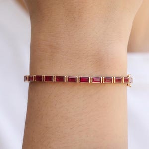 Puede incluir: Una pulsera de tono dorado que presenta una hilera continua de piedras rectangulares de color rojo intenso con talla baguette engastadas en un marco de metal. La pulsera rodea la muñeca y se asegura con un pequeño cierre integrado lateral.