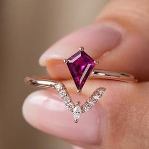 May include: A rose gold ring featuring a kite-shaped, deep pink gemstone. Beneath it, a second ring with a curved design is embellished with small, clear gemstones. The rings are held in a person's hand.