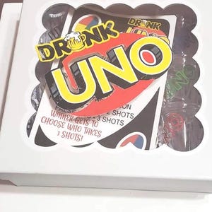 May include: A white box containing a card game called "Drunk Uno." The game's logo features the words "Drunk Uno" in yellow and red, with a beer mug graphic. The box also includes shot glasses and game instructions.
