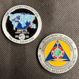 May include: Two circular metal challenge coins. One features a blue world map, the text "YOU CAN RUN BUT YOU CAN'T HIDE," "BIRS 79," "JAN 6 - MAR 12," "20 20," and "THE HUNT BEGINS HERE." The other displays the Department of Justice seal with a globe.