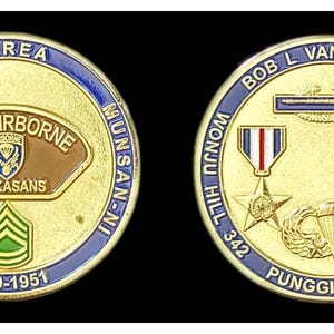 May include: A gold-coloured commemorative coin with blue and white trim. The coin features text and military insignia, including the words "KOREA," "187th AIRBORNE," and "RAKKASANS." The coin also has the text "1950-1951."