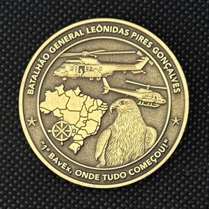 May include: A bronze-colored coin featuring helicopters, a map, and an eagle. The coin has text around the edge that reads "BATALH&Atilde;O GENERAL LEONIDAS PIRES GON&Ccedil;ALVES" and "1&deg; BAvEx, ONDE TUDO COME&Ccedil;OU!"