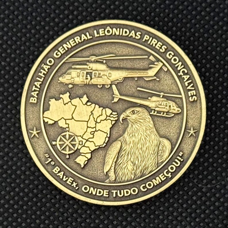 May include: A bronze-colored coin featuring helicopters, a map, and an eagle. The coin has text around the edge that reads "BATALH&Atilde;O GENERAL LEONIDAS PIRES GON&Ccedil;ALVES" and "1&deg; BAvEx, ONDE TUDO COME&Ccedil;OU!"