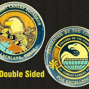 May include: Double-sided coin with the text "3D Combat Camera Squadron" and "JBSA-Lackland, Texas". One side features an eagle with a camera and weapon. The other side reads "Presented by the Commander".