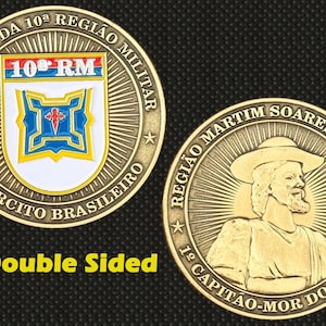 以下が含まれることがあります： 両面金色のコイン。片面には「10ª RM」と「EXERCITO BRASILEIRO」の文字が入った軍事エンブレムが描かれています。もう片面には、帽子をかぶった男性と「REGIÃO MARTIM SOARES MORENO」の文字が描かれています。
