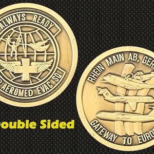 Puede incluir: Una moneda dorada de doble cara. Un lado presenta el texto "ALWAYS READY" y "2ND AEROMED EVAC SQ" alrededor de un globo y una cruz. El otro lado muestra tres aviones y el texto "RHEIN MAIN AB, GERMANY" y "GATEWAY TO EUROPE".