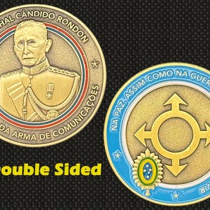 May include: Double-sided gold-colored coin featuring a portrait of Marechal Cândido Rondon and text in Portuguese. The reverse side has a gold design with arrows and text in Portuguese. The coin is set against a black background.