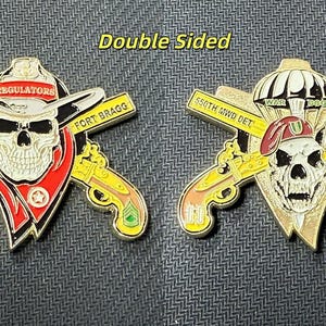 May include: Double-sided military challenge coin featuring a skull with a cowboy hat and crossed pistols. One side reads "Regulators" and "Fort Bragg". The other side features a skull with a beret and reads "War Dogs" and "Fort Liberty".