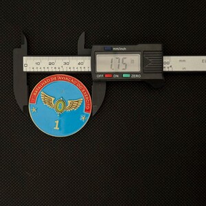 May include: A blue, red, and gold military coin, approximately 1.75 inches in diameter, is measured with a digital caliper. The coin features a winged emblem and the text "1&deg; BATALH&Atilde;O DE AVIA&Ccedil;&Atilde;O DO EX&Eacute;RCITO".