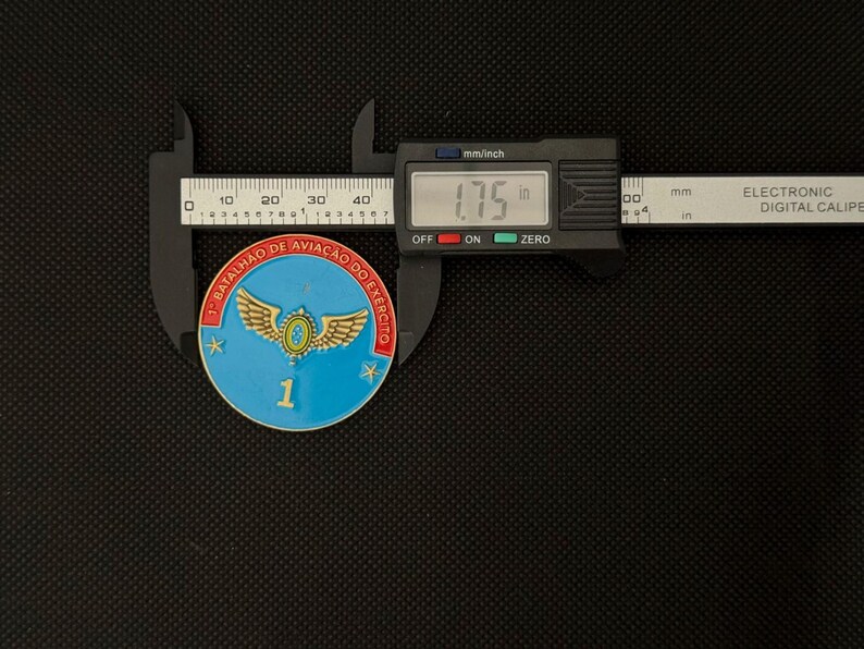 May include: A blue, red, and gold military coin, approximately 1.75 inches in diameter, is measured with a digital caliper. The coin features a winged emblem and the text "1&deg; BATALH&Atilde;O DE AVIA&Ccedil;&Atilde;O DO EX&Eacute;RCITO".