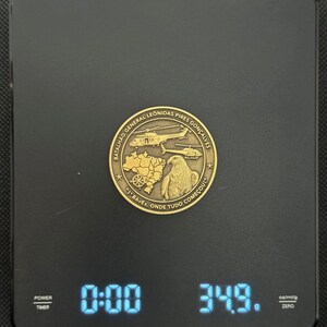 May include: A gold-colored coin with text and images of helicopters, a map, and an eagle is displayed on a black digital scale. The scale reads 34.9 grams. The coin has the text "Batalh&atilde;o General Leonidas Pires Gon&ccedil;alves" and "1 BAVEx, Onde Tudo Come&ccedil;ou!"