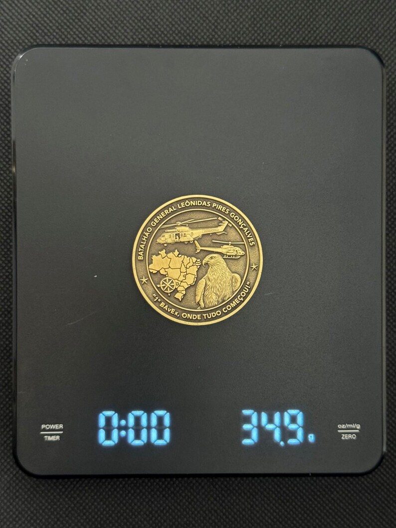 May include: A gold-colored coin with text and images of helicopters, a map, and an eagle is displayed on a black digital scale. The scale reads 34.9 grams. The coin has the text "Batalh&atilde;o General Leonidas Pires Gon&ccedil;alves" and "1 BAVEx, Onde Tudo Come&ccedil;ou!"
