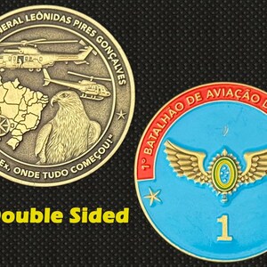 May include: Double-sided commemorative coin. One side features helicopters, a map, and an eagle, with text in Portuguese. The other side is blue with a winged emblem and the number 1. The text "Double Sided" is also present.