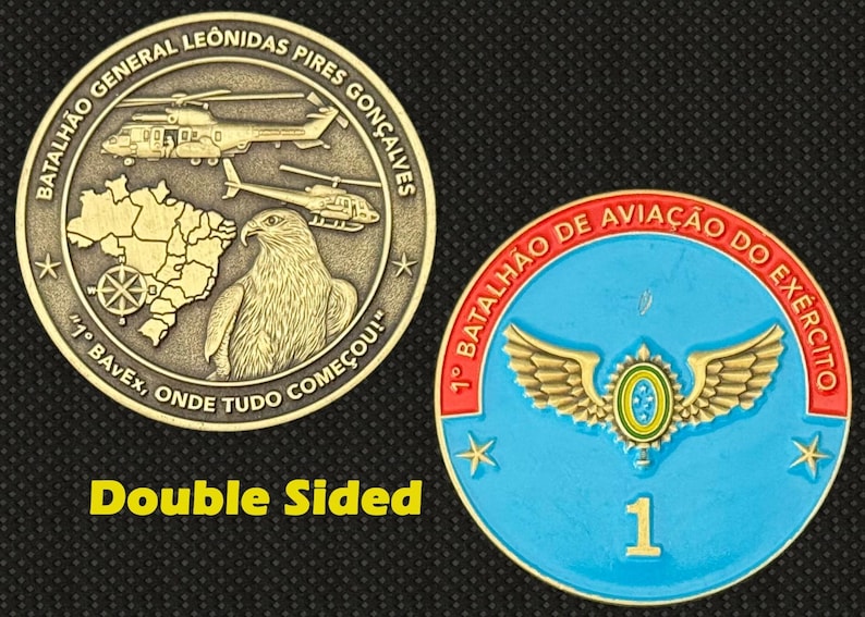 May include: Double-sided commemorative coin. One side features helicopters, a map, and an eagle, with text in Portuguese. The other side is blue with a winged emblem and the number 1. The text "Double Sided" is also present.