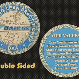 May include: Double-sided coin with blue and silver accents. One side features the text "ENGINEERING LEAN PRACTITIONER" and the Daikin logo. The other side lists "OUR VALUES". The coin is a collectible item.
