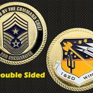 May include: Double-sided gold coin with detailed designs. One side shows a military insignia with stars and stripes, the other side displays a shield with a desert landscape and the text "162D WING". The coin is presented by the Command Chief.
