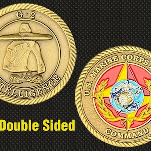 Może przedstawiać: Dwustronna moneta w kolorze złotym. Jedna strona przedstawia postać z szerokim kapeluszem i napisem "G-2 INTELLIGENCE". Druga strona przedstawia emblemat U.S. Marine Corps Forces z tekstem. Moneta ma obramowanie w kształcie liny.