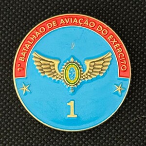 May include: A round, blue military emblem with gold trim and text. The emblem features a central design of wings and a circular emblem, with the number "1" in gold at the bottom. The text "1&deg; BATALH&Atilde;O DE AVIA&Ccedil;&Atilde;O DO EX&Eacute;RCITO" is in red.
