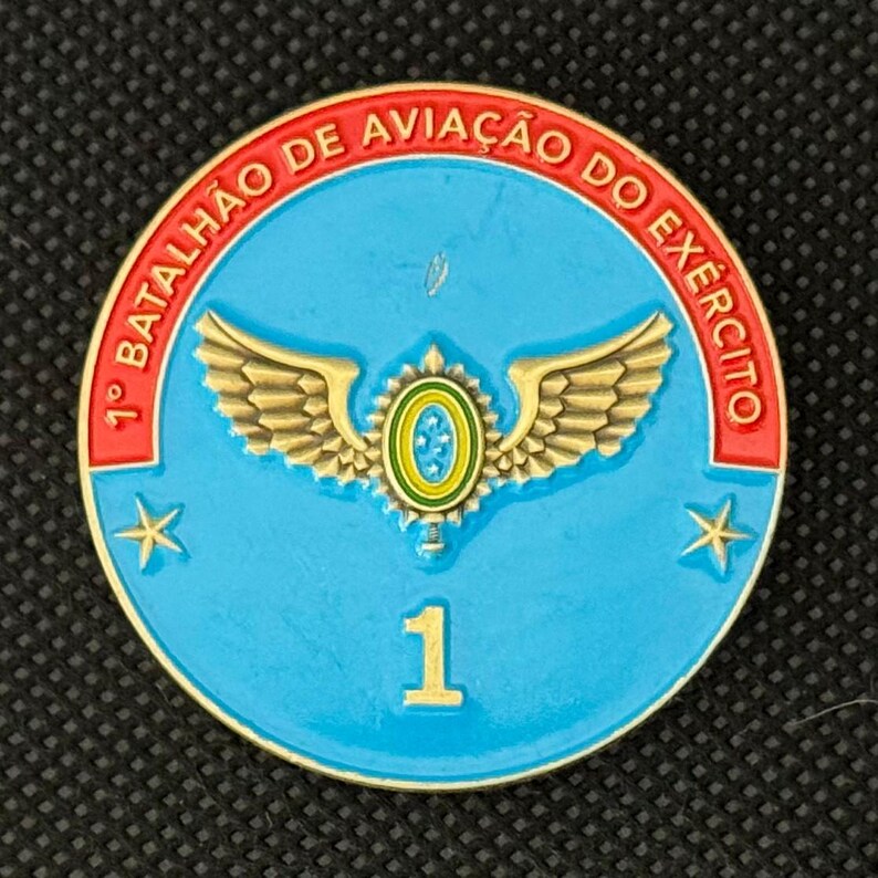 May include: A round, blue military emblem with gold trim and text. The emblem features a central design of wings and a circular emblem, with the number "1" in gold at the bottom. The text "1&deg; BATALH&Atilde;O DE AVIA&Ccedil;&Atilde;O DO EX&Eacute;RCITO" is in red.