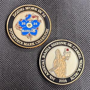May include: Two circular gold-toned metal challenge coins with black enamel borders. One coin features a blue flower emblem with a dragon, the text "DOING MORE IN 24" and "POTENTATE MARK CHARLTON". The other shows a camel and the text "5TH ANNUAL BAHIA SHRINE 5K CAMEL WALK/RUN 2024".