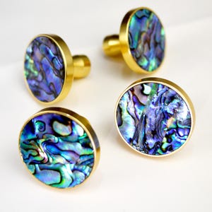 May include: Four decorative cabinet knobs with iridescent abalone shell centers and gold-toned metal rims and stems. The abalone shell displays shades of blue, green, and purple. The knobs are arranged on a white surface.