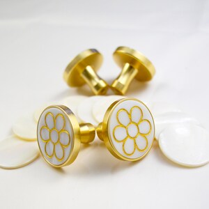 May include: Four gold-toned cufflinks with a white and gold floral design. The cufflinks have a round face with a white background and a gold flower design. The cufflinks are surrounded by white, oval-shaped objects.