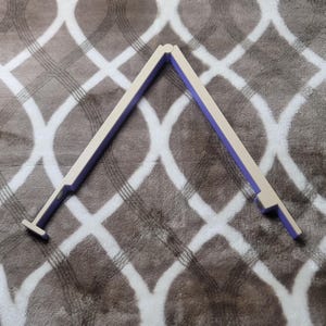May include: A white and purple adjustable tool, possibly for measuring or marking. The tool is in an open, angled position, with a T-shaped end on one side. The background is a patterned, soft, gray surface.