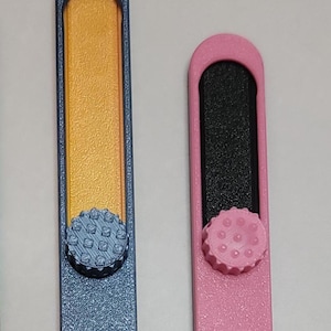 May include: Two nail files. One is blue with a gold center and a textured knob. The other is pink with a black center and a pink textured knob. Both have a rectangular shape with a rounded top.