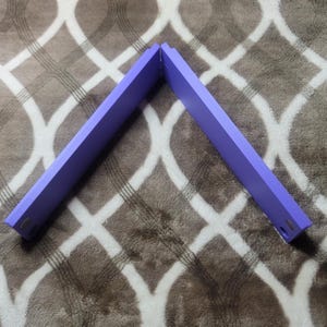 May include: Two lavender-colored wooden shelves joined at a 90-degree angle, forming an L-shape. Each shelf has a rectangular profile with rounded corners and two small holes at the end. The shelves are set against a patterned, brown and white background.