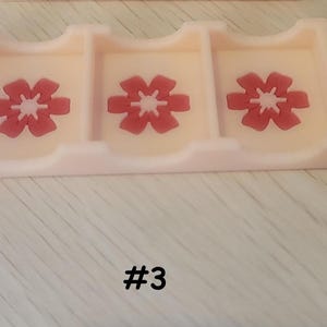 May include: A light pink card holder with three compartments, each featuring a red flower design. The number "#3" is printed in black below the holder. The holder is made of a smooth, solid material.