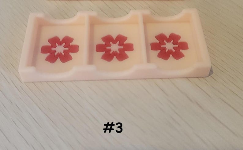 May include: A light pink card holder with three compartments, each featuring a red flower design. The number "#3" is printed in black below the holder. The holder is made of a smooth, solid material.