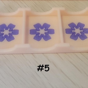 May include: A light pink rectangular tray with three compartments, each decorated with a purple flower design. The number "#5" is printed in black at the bottom of the tray. The tray is likely used for organizing small items.
