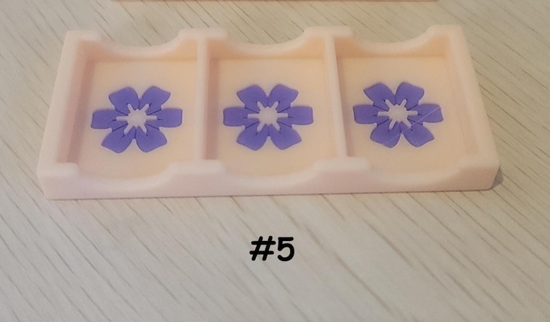 May include: A light pink rectangular tray with three compartments, each decorated with a purple flower design. The number "#5" is printed in black at the bottom of the tray. The tray is likely used for organizing small items.