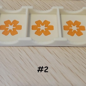 May include: A white plastic organizer with three compartments, each featuring an orange flower design. The number "#2" is printed in black at the bottom. The organizer sits on a light-colored wooden surface.