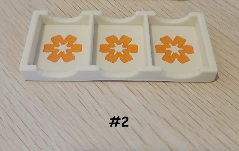 May include: A white plastic organizer with three compartments, each featuring an orange flower design. The number "#2" is printed in black at the bottom. The organizer sits on a light-colored wooden surface.