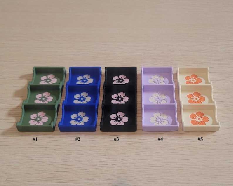 May include: Five miniature, three-tiered display stands in various colors: green, blue, black, lavender, and cream. Each stand features a floral design on each tier. The stands are numbered from one to five.