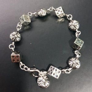 May include: Silver bracelet with dice-shaped charms and flower-shaped beads. The dice have black markings. The bracelet is connected by small chain links and has a clasp closure.