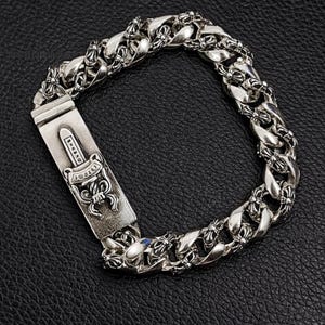 Könnte beinhalten: Silberfarbenes Armband mit einem dicken Panzerketten-Design. Der Verschluss hat eine rechteckige Platte mit einem Dolch- und Schriftrollen-Design. Das Armband wird auf einer schwarzen, strukturierten Oberfläche gezeigt.