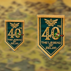 Puede incluir: Dos parches bordados en forma de escudo en verde oscuro y dorado. Cada parche presenta el texto 40TH THE LEGEND OF ZELDA, un emblema de la Trifuerza dorada y un pequeño gráfico de espada verde y roja integrado en el número cero del bordado.