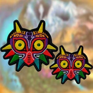 Peut inclure: Deux écussons brodés représentant un masque stylisé avec une bordure noire. Le masque présente de grands yeux jaunes, une section supérieure violette, une zone centrale rouge et des pointes rayonnantes jaunes, rouges, vertes et bleu sarcelle.